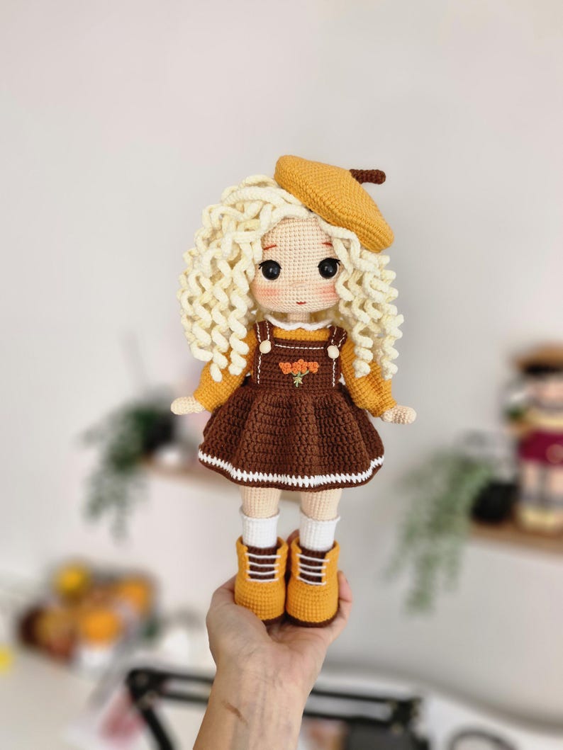 May include: A crocheted doll with blonde curly hair, wearing a brown dress with a white trim, a yellow beret, and yellow boots.