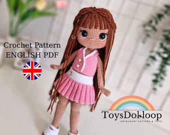 Puede incluir: Una muñeca de ganchillo hecha a mano con cabello castaño y un atuendo rosa. La muñeca tiene una parte superior rosa con ribete blanco y una falda plisada a juego. Lleva zapatos blancos y tiene ojos grandes y expresivos. El texto "Crochet Pattern ENGLISH PDF" es visible.