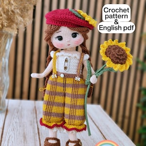May include: A handcrafted crochet doll wearing a red beret with a sunflower detail, a white top, yellow and brown striped dungarees, and brown shoes. The doll holds a crochet sunflower. The image includes the text "Crochet pattern & English pdf".