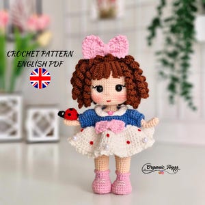 May include: Crochet pattern for a doll with brown curly hair, a pink bow, a blue and white dress, and pink shoes. The doll is holding a ladybug. The pattern is available in English PDF format.