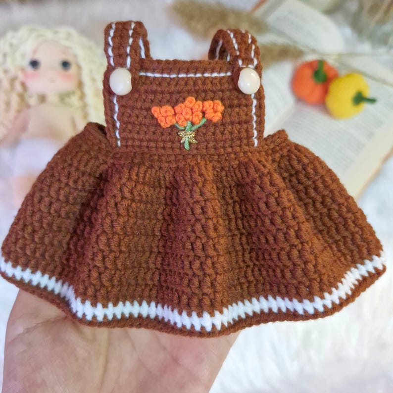 May include: A brown crocheted dress with white trim and an orange flower design. The dress has two white buttons on the straps.