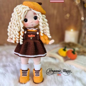 Pode incluir: Padr&atilde;o de boneca de croch&ecirc; em ingl&ecirc;s. A boneca est&aacute; vestindo um vestido marrom com uma borda branca, um su&eacute;ter amarelo, um bon&eacute; amarelo e sapatos amarelos. A boneca tem cabelo loiro encaracolado e olhos castanhos. O fundo &eacute; uma superf&iacute;cie de madeira com um livro e uma vela.