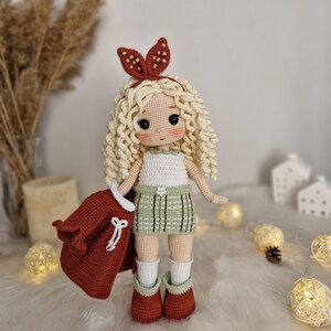 Op de afbeelding: Een gehaakte pop met blond krullend haar, die een wit tanktopje, een groene geruite rok, een rood jasje en bruine laarzen draagt. De pop heeft een rood hoofdband met witte stippen.