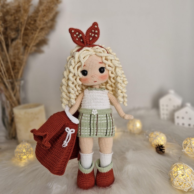 Op de afbeelding: Een gehaakte pop met blond krullend haar, die een wit tanktopje, een groene geruite rok, een rood jasje en bruine laarzen draagt. De pop heeft een rood hoofdband met witte stippen.