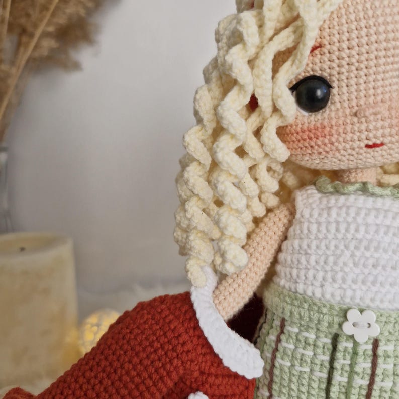 Pode incluir: Uma boneca de croch&ecirc; com cabelo comprido e encaracolado loiro, vestindo um top branco sem mangas e uma saia verde com uma flor branca. A boneca est&aacute; segurando um objeto vermelho de croch&ecirc;.