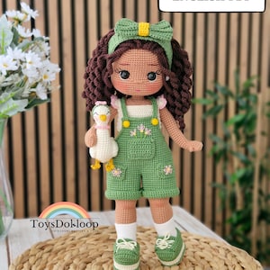 Pode incluir: Uma boneca de crochê com cabelo castanho escuro encaracolado, usa uma faixa verde com laço, um top branco e jardineiras verdes com bordados florais. A boneca segura um pequeno pato branco e usa meias brancas com tênis verdes. Texto: Crochet Pattern ENGLISH PDF.