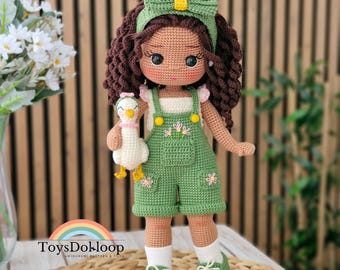May include: A crochet doll with dark brown curly hair, wearing a green headband with a bow, a white top, and green overalls with floral embroidery. The doll holds a small white duck toy and wears white socks with green sneakers. Text reads: Crochet Pattern ENGLISH PDF.