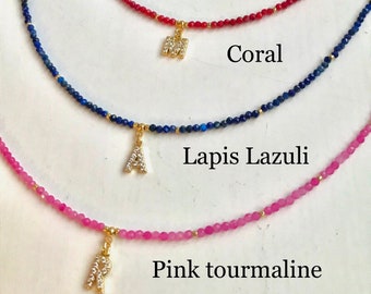 May include: A collection of gemstone necklaces in yellow jade, onyx, coral, lapis lazuli, and pink tourmaline. Each necklace features a different colored bead strand and a gold-colored initial pendant. The names of the gemstones are labeled.