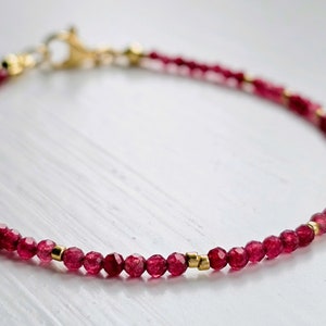 Könnte beinhalten: Ein zartes Armband mit kleinen, facettierten, tiefroten Edelsteinen. Das Armband ist mit kleinen goldfarbenen Perlen und einem goldfarbenen Verschluss versehen. Der Schmuck wird auf einer weißen Oberfläche präsentiert.
