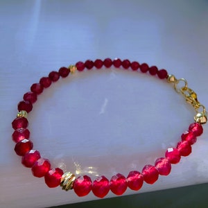 May include: A red beaded bracelet with gold-coloured accents. The bracelet features faceted red beads strung together with gold-coloured spacers and a gold-coloured clasp. The bracelet is a delicate piece of jewellery.