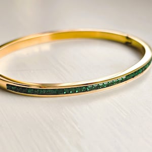 May include: A gold-colored bangle bracelet with a row of small, square, emerald-green gemstones set along the front. The bracelet has a smooth, polished finish and a simple, elegant design. This piece of jewelry is suitable for everyday wear or special occasions.