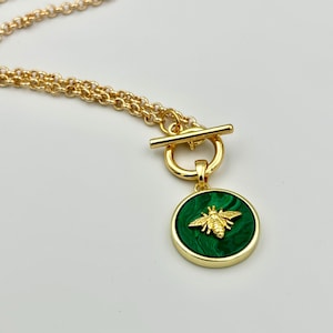Op de afbeelding: Goudkleurige ketting met een ronde hanger met een groen gemarmerd ontwerp en een gouden bijen-embleem. De ketting heeft een tuimelsluiting en een fijne ketting, perfect om een vleugje elegantie toe te voegen.