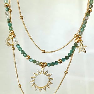 Op de afbeelding: Een gelaagde ketting set met gouden kettingen en kralen strengen. De kettingen bevatten een maan hanger, een zon hanger en een ster hanger, allemaal versierd met heldere kristallen. De kralen zijn goud en groen.