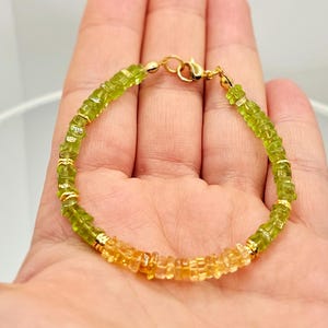 May include: A beaded bracelet featuring a gradient of green and yellow gemstones. The bracelet is strung on a gold chain and has a gold clasp. The gemstones are small and evenly spaced, creating a delicate and elegant piece of jewelry.