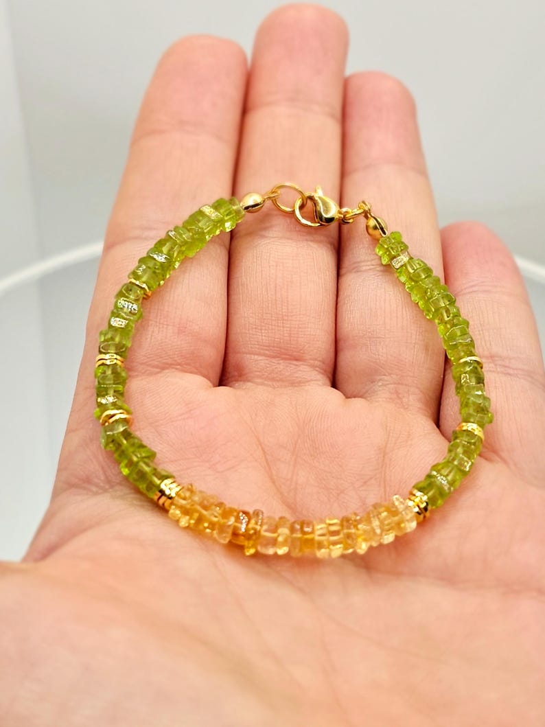 May include: A beaded bracelet featuring a gradient of green and yellow gemstones. The bracelet is strung on a gold chain and has a gold clasp. The gemstones are small and evenly spaced, creating a delicate and elegant piece of jewelry.