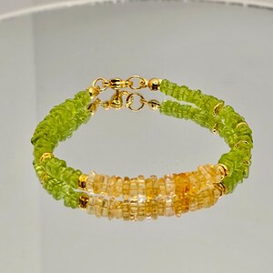 May include: A double-strand bracelet featuring light green and yellow-orange gemstones. The bracelet has gold-colored clasps and small gold beads. The gemstones are small and square-cut, creating a textured appearance. The bracelet is displayed on a reflective surface.