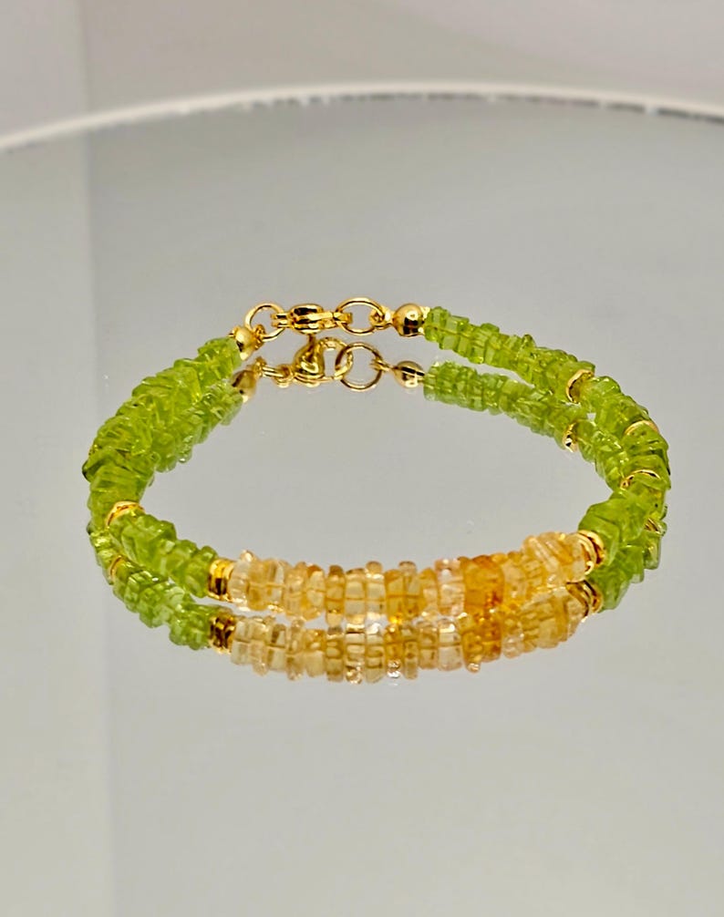 May include: A double-strand bracelet featuring light green and yellow-orange gemstones. The bracelet has gold-colored clasps and small gold beads. The gemstones are small and square-cut, creating a textured appearance. The bracelet is displayed on a reflective surface.