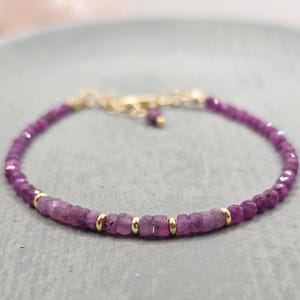 May include: A delicate bracelet featuring small, faceted, deep pink gemstones interspersed with gold-coloured beads. The bracelet has a gold-coloured chain and clasp, creating a refined and elegant piece of jewellery.