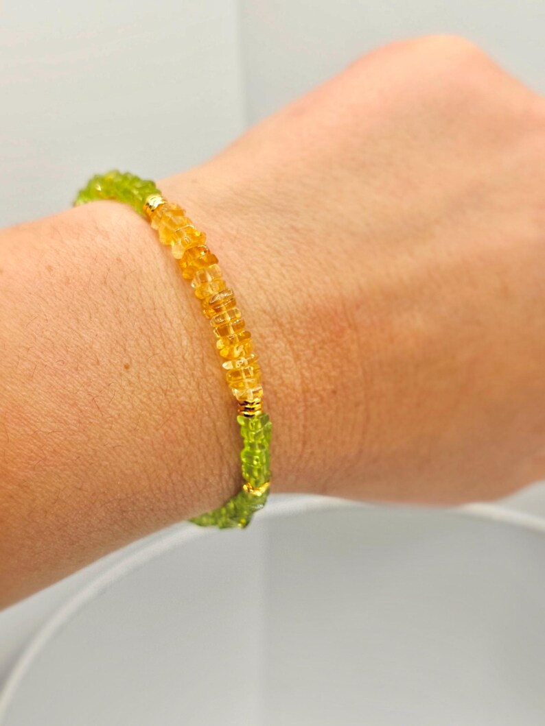 May include: A beaded bracelet featuring a combination of green and yellow gemstones. The bracelet is strung with small, faceted beads in shades of yellow and green, with gold-colored accents. The bracelet is worn on a wrist.