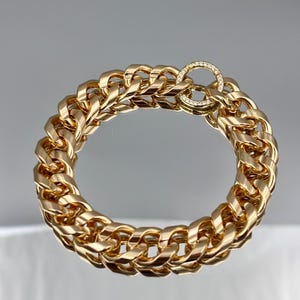 May include: A gold-colored chain bracelet with a thick, curb link design. The bracelet features a circular clasp adorned with small, clear gemstones. The jewelry is displayed on a reflective surface, highlighting its polished finish.