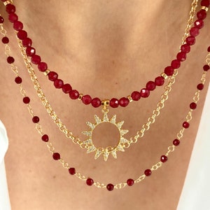 May include: A multi-strand necklace with red beads and gold chains. The top and bottom strands have small, faceted, red beads. The middle strand features a gold chain with a sunburst pendant. The pendant is encrusted with small, clear crystals.