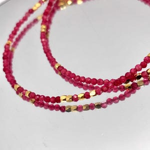 Può includere: Primo piano di una delicata collana con piccole perline rotonde rosso intenso intervallate da perline dorate di forma geometrica. La collana è esposta su una superficie riflettente, creando un effetto specchio. Il gioiello è probabilmente un accessorio di moda.