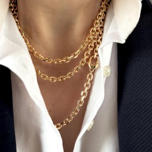 May include: Three gold-coloured chain necklaces of varying lengths. The chains have an oval link design and are layered over a white collared shirt and a navy blue blazer. The necklaces are secured with a lobster clasp.
