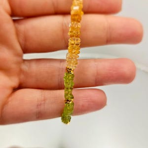 May include: A beaded bracelet featuring a mix of green and yellow-orange gemstones. The bracelet is strung together with gold-colored accents. The gemstones are small and irregularly shaped, creating a textured appearance. The bracelet is held up against a light background.