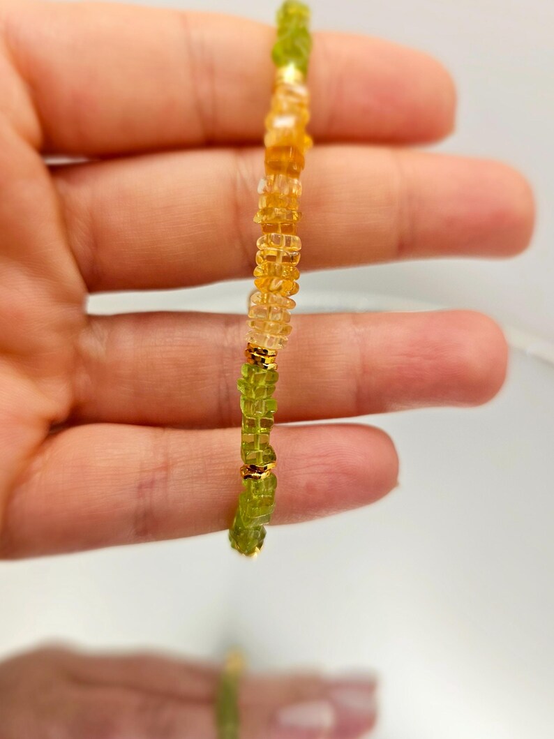 May include: A beaded bracelet featuring a mix of green and yellow-orange gemstones. The bracelet is strung together with gold-colored accents. The gemstones are small and irregularly shaped, creating a textured appearance. The bracelet is held up against a light background.