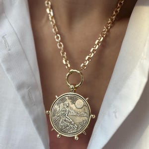 May include: A gold-toned necklace featuring a thick, round link chain and a large, circular pendant. The pendant displays a detailed embossed scene of a figure sitting by a body of water under a moon, with small decorative spikes around the edge.