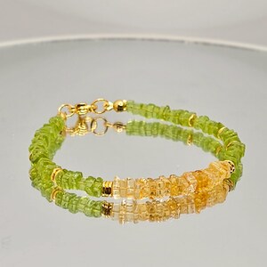 May include: A delicate bracelet featuring small, faceted green and yellow gemstones. The bracelet is strung on a gold chain with a gold clasp. The gemstones are arranged in a repeating pattern, creating a subtle sparkle.