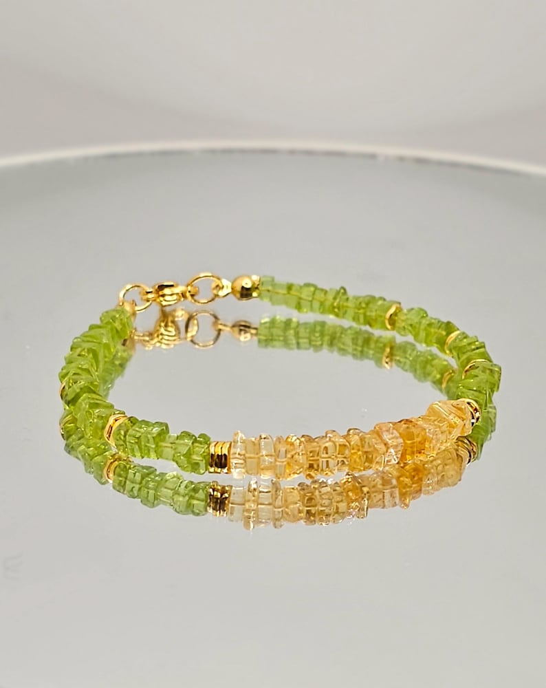 May include: A delicate bracelet featuring small, faceted green and yellow gemstones. The bracelet is strung on a gold chain with a gold clasp. The gemstones are arranged in a repeating pattern, creating a subtle sparkle.