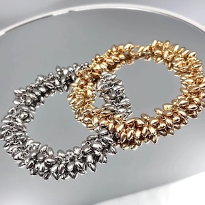 May include: Two beaded bracelets, one silver and one gold, are shown on a reflective surface. The bracelets are circular, with each bead shaped like a small leaf. The gold bracelet is next to the silver bracelet.