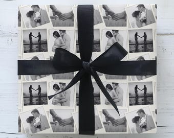May include: Square gift box wrapped in paper printed with black and white photos of couples. A black satin ribbon is tied around the box with a large bow. The box is on a white wooden surface.