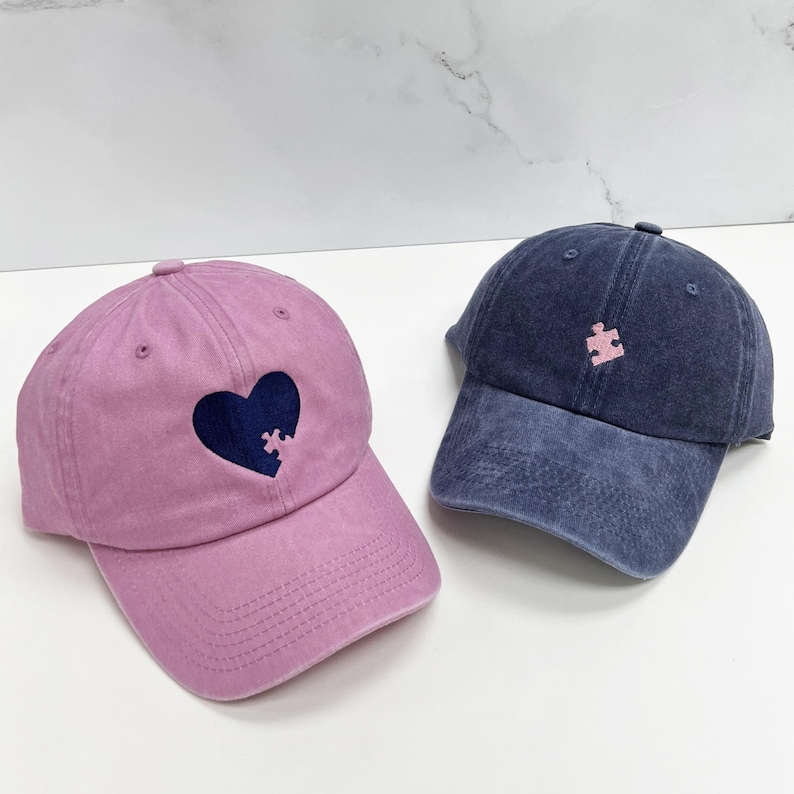 May include: Two denim baseball caps, one pink and one blue, with embroidered puzzle piece designs. The pink cap has a heart shape with a blue puzzle piece inside.