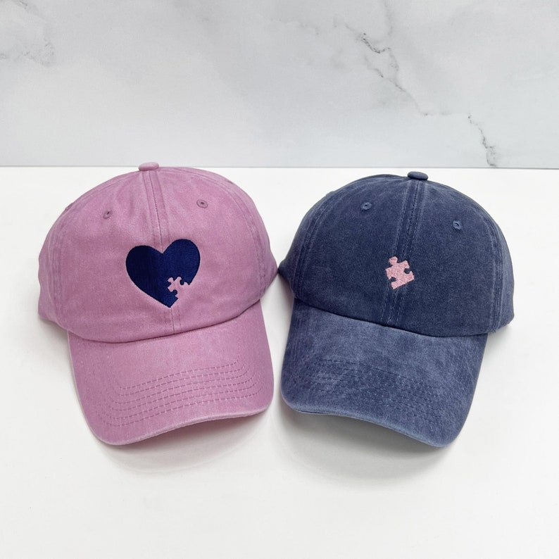 May include: Two baseball caps, one pink and one blue, with embroidered designs. The pink cap has a heart with a puzzle piece missing, and the blue cap has a puzzle piece in the shape of a heart.