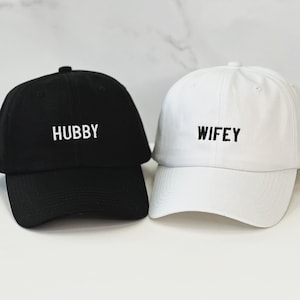 Op de afbeelding: Twee baseballpetten, een zwarte met het woord "HUBBY" geborduurd in wit, en een witte met het woord "WIFEY" geborduurd in zwart.