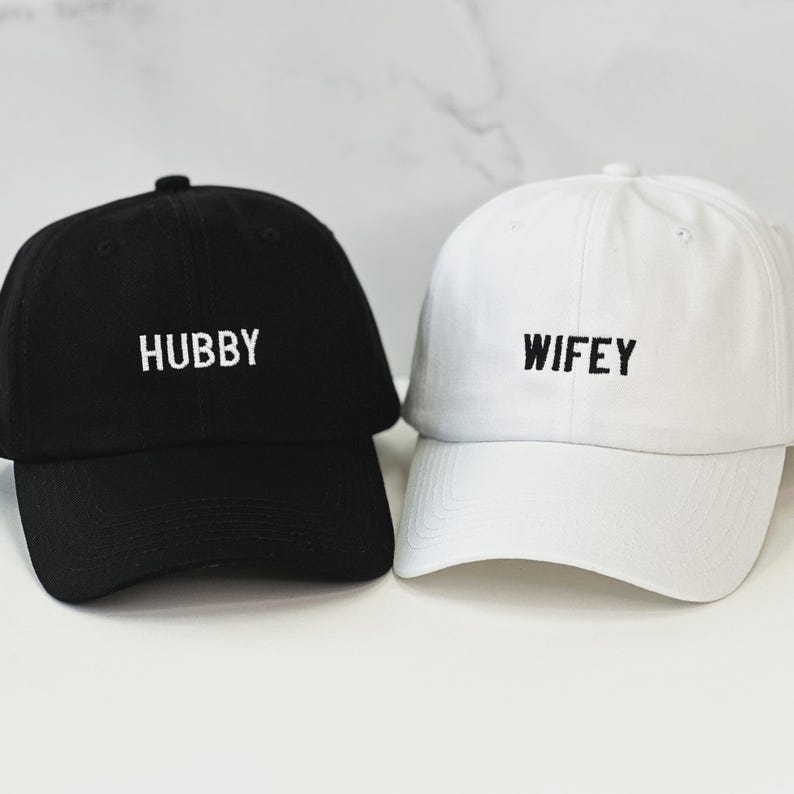 Op de afbeelding: Twee baseballpetten, een zwarte met het woord "HUBBY" geborduurd in wit, en een witte met het woord "WIFEY" geborduurd in zwart.