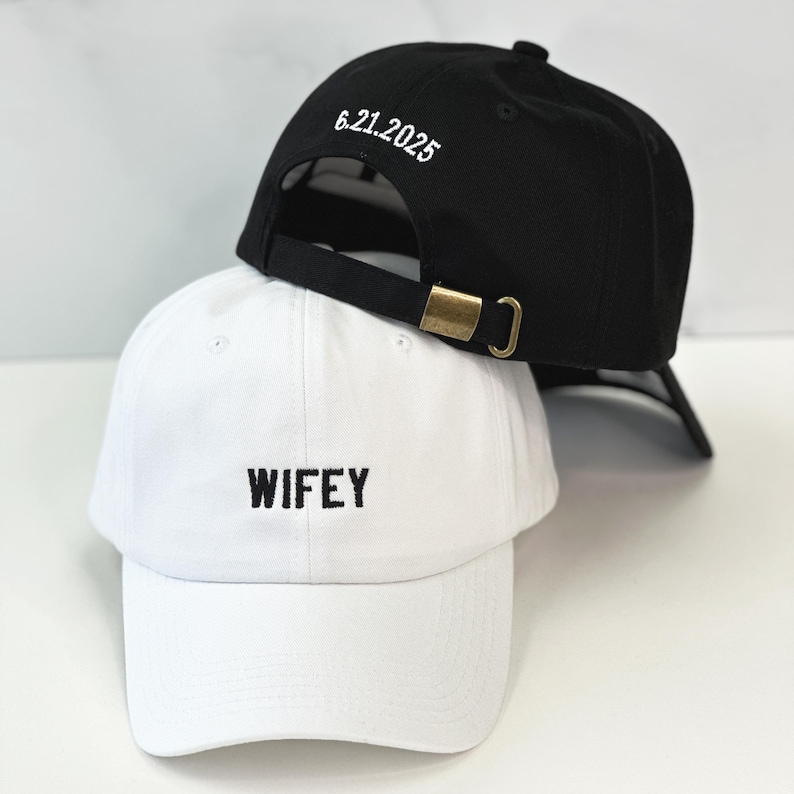 Op de afbeelding: Twee baseballpetten, een zwarte en een witte. De zwarte pet heeft de datum "6.21.2025" erop geborduurd. De witte pet heeft het woord "WIFEY" erop geborduurd.