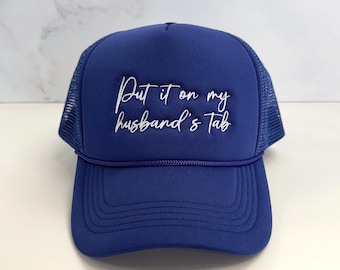 May include: A royal blue trucker hat with a white mesh back and a curved brim. The hat features the embroidered phrase "Put it on my husband's Tab" in a cursive font across the front. A casual accessory.