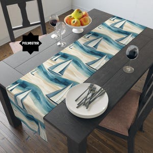 May include: A table runner with a nautical design featuring sailboats on a watercolor-style ocean. The runner is primarily blue, beige, and white, with the word "POLYESTER" in a black starburst shape. The runner is on a dark wooden table.