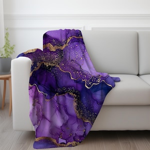Pode incluir: Cobertor abstrato roxo e dourado com um design de mármore. O cobertor apresenta redemoinhos de tinta roxa e dourada com um efeito sutil de brilho.