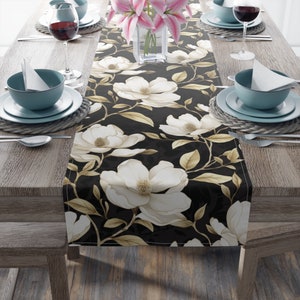 May include: A black table runner with a white floral pattern. The runner features large white magnolia flowers with gold accents and brown leaves on a black background.