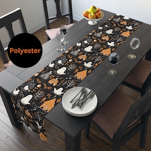 May include: Black polyester table runner with a Halloween-themed pattern. The design features white ghosts, orange houses, and autumn foliage. The text "Polyester" is in a black circle. The runner is on a dark wooden table.