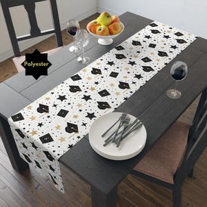 May include: White table runner with a black and gold graduation cap and star pattern. The text "Polyester" is visible in the image.
