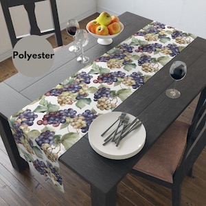 May include: A white table runner with a watercolor design of grapes and leaves. The runner is on a dark wood table with two wine glasses and a bowl of apples. The table runner is made of polyester.