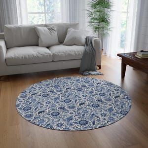May include: A round blue and white floral patterned rug with a white background and blue floral designs. The rug is in a living room setting with a white couch and a wooden coffee table.
