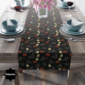 Puede incluir: Camino de mesa negro con temática de Halloween con un patrón repetido de casas encantadas, fantasmas y calabazas. El camino de mesa está hecho de poliéster y está colocado sobre una mesa de madera con cubiertos y un jarrón con flores rosas.