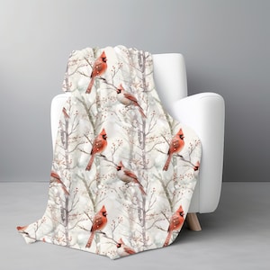 May include: A cozy throw blanket draped over a white armchair. The blanket features a repeating pattern of red cardinal birds perched on bare tree branches with red berries, set against a snowy, light gray background. The blanket is made of soft material.