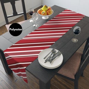 Puede incluir: Un camino de mesa de poliéster a rayas rojas y blancas. El camino de mesa está colocado sobre una mesa de madera oscura, con un cuenco blanco de manzanas, copas de vino y un plato con cubiertos.